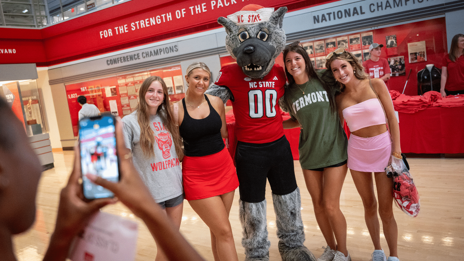 A person takes a photo of four students with Mr. Wuf standing in Reynolds Coliseum.